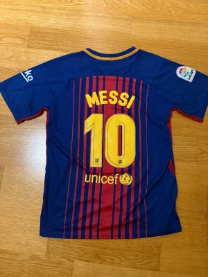FC Barcelona Messi matchtröja - Säljer en officiell FC Barcelona matchtröja från Nike med Messi 10 på ryggen. Tröjan är blå med röda vertikala ränder, gula detaljer och klubbmärke på bröstet. Materialet är lätt och andas, perfekt för fotbollsträning eller att bära på läktaren.