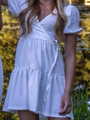 Vit studentklänning  - Söt vit kortklänning med omlottmodell och puffärmar. Klänningen har knyt i midjan och volang nedtill. Perfekt inför studenten💕