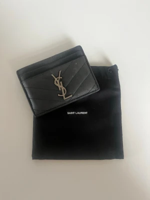 Svart kortfodral från Saint Laurent - Snyggt svart kortfodral i läder från Saint Laurent med silverfärgad YSL-logga framtill. Fodralet har flera kortfack och en stilren, quiltad design. Perfekt accessoar för dig som gillar lyxiga detaljer och minimalistisk stil.