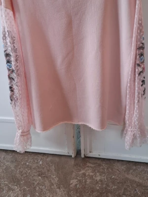 Rosa blus med broderade ärmar - Supersöt ljusrosa blus med transparenta, prickiga långärmar i mesh och broderade blommor i rosa, blått och gult. Ärmarna har volang vid ärmslutet och ger en romantisk vibe. Perfekt för dig som gillar detaljer och pastellfärger.