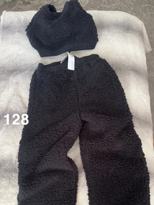 Svart teddyset med topp och byxor - Mysigt svart teddyset med croppad topp och matchande byxor. Båda delarna har bred resår med texten 'STAY COOL'. Perfekt för att chilla hemma eller som ett trendigt loungeplagg. Materialet är fluffigt och mjukt för extra komfort.🌸 Ny med lapp. Prislappen sitter inte fast i båda plaggen. ( påverkar inte funktionen) Storlek 128