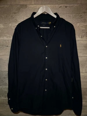 Mörkblå skjorta från Polo Ralph Lauren - Klassisk mörkblå skjorta från Polo Ralph Lauren med knappslå och ikoniska broderade loggan på bröstet. Skjortan har lång ärm, button-down krage och vita knappar. Perfekt till jeans eller chinos för en clean look.