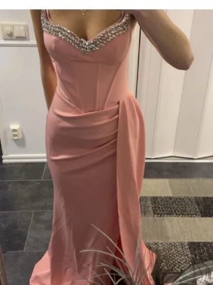 Bal klänning - Elegant långklänning i rosa med figurnära passform och vackra pärldetaljer längs urringningen. Klänningen har en draperad detalj vid höften och markerad midja för en snygg siluett. Perfekt för speciella tillfällen. Aldrig använd 