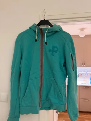 Turkos hoodie med dragkedja från Peak Performance - Turkos hoodie från Pelle P med dragkedja framtill, huva med dragsko och broderad logga på bröstet. Fickor på sidorna och en extra ficka med dragkedja på ärmen. Perfekt för en avslappnad och sportig stil.