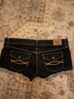 Mörkblå jeansshorts Abercrombie & Fitch - Snygga mörkblå jeansshorts från Abercrombie & Fitch med råa kanter och dekorativa bakfickor med gul söm. Klassisk femficksmodell och läderpatch med logga bak i midjan. Perfekta för sommaren!
