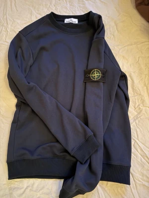 Mörkblå sweatshirt från Stone Island - Mörkblå crewneck sweatshirt från Stone Island med den ikoniska kompasspatchen på ärmen. Tröjan har ribbade muddar vid ärmslut och nederkant samt en klassisk rund halsringning. Perfekt för en stilren och avslappnad look.