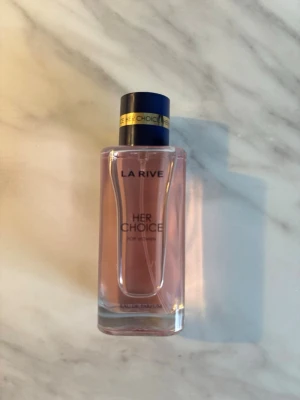 La Rive Her Choice parfym - La Rive Her Choice är en parfym för kvinnor med en elegant, rektangulär glasflaska och ljusrosa doftvätska. Flaskan har en svart kork med gulddetalj och text. Perfekt för dig som gillar stilrena och feminina dofter.
