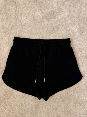 Svarta shorts med snörning - Svarta shorts med elastisk midja och snörning framtill. Klassisk sportig modell med korta ben och mjukt material, perfekta för avslappnade dagar eller träning.