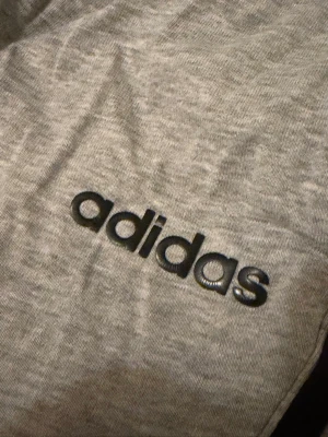 ADIDAS mjukisbyxor med svarta snören - Säljer ett par grå mjukisbyxor med svarta dragsnören i midjan och elastisk resår. Byxorna har en avslappnad passform och är tillverkade i mjukt material, perfekt för chill dagar. Diskreta detaljer och klassisk look.