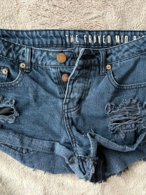 Blå jeansshorts med fransar - PRIS GÅR ATT DISKUTERA. Säljer ett par blå jeansshorts från The Frayed med råa kanter och slitna detaljer framtill och baktill. Shortsen har midwaist passform, två knappar i midjan och klassiska fickor både fram och bak. Perfekta för sommaren!