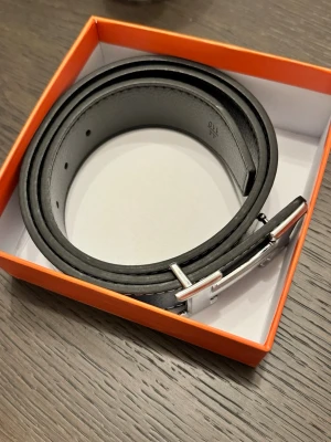 Svart skärp med silverfärgat spänne - Stilrent svart bälte med klassiskt silverfärgat spänne. Bältet har en slät design och är tillverkat i skinn, perfekt för att lyfta vilken outfit som helst. Levereras i en snygg orange ask.