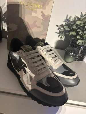 Valentino Garavani camo sneakers - Snygga sneakers från Valentino Garavani med svart, grå och beige camo-mönster. Skorna har vita snören, robust svart sula med mönster och detaljer i både läder och textil. Perfekta för dig som vill sticka ut med exklusiv streetstyle.