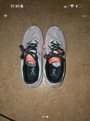 On Cloudmonster vita sneakers - Säljer ett par On Cloudmonster sneakers i vitt med svarta och orange detaljer. Skorna har svart/vita snören, tjock sula med unik design och meshmaterial för bra ventilation. Perfekta för dig som gillar sportig och modern stil.