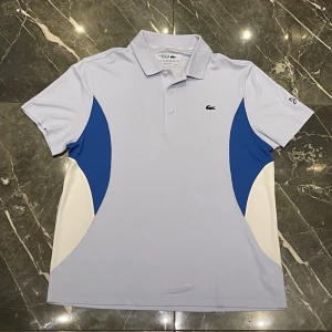 Ljusblå pikétröja från Lacoste - Snygg ljusblå pikétröja från Lacoste med klassisk krage och knappar framtill. Tröjan har vita och blå paneler på sidorna samt den ikoniska krokodilloggan på bröstet. Perfekt för en sportig och stilren look.