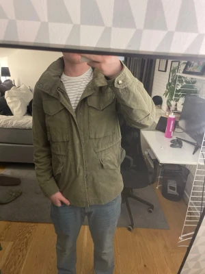 Olivgrön vindjacka med huva - Säljer en olivgrön vindjacka med huva och flera praktiska fickor framtill. Jackan har dragkedja och tryckknappar, samt justerbar midja för skön passform. Perfekt för dig som vill ha en stilren och funktionell jacka till vardags. Materialet känns slitstarkt och lätt. Sälsynt 