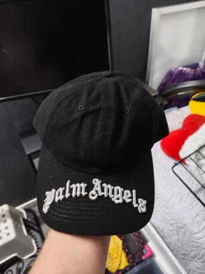 Svart Palm Angels keps - Säljer en svart keps från Palm Angels med böjd skärm och vit broderad logga framtill. Klassisk sexpanel-design och justerbar passform bak. Tillverkad i bomull för en bekväm känsla och streetwear-vibe.