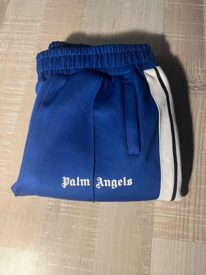 Blåa Palm Angels mjukisbyxor - Säljer ett par blåa mjukisbyxor från Palm Angels med vita och svarta ränder längs sidorna. Byxorna har resår i midjan och fickor på sidorna. Tillverkade i polyester för en skön och sportig känsla. Perfekta för en avslappnad och trendig look. Stolek S men passar xs!  Skicka bud
