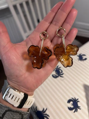 Bruna blommigad örhängen  - örhängen med  blommor i brun och bärnstensfärgad plast. Hängande modell med guldfärgade detaljer och ringar. Perfekta för dig som vill sticka ut med en cool accessoar. Blommorna har en snygg, glansig finish och organisk form.