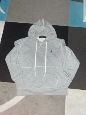 Grå hoodie från Polo Ralph Lauren - Snygg grå hoodie från Polo Ralph Lauren med klassisk huva och vita dragsnören. Har en liten blå logga broderad på bröstet och stor magficka framtill. Tillverkad i mjuk bomullsblandning som är perfekt för chill dagar.
