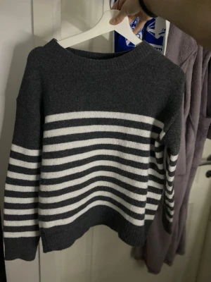 Mörkgrå randig stickad tröja - Mysig mörkgrå stickad tröja med vita horisontella ränder på både kropp och ärmar. Tröjan har en klassisk rund halsringning och ribbade muddar vid ärmslut och nederkant. Perfekt för kyliga dagar och enkel att matcha.