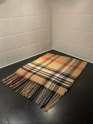 Beige rutig halsduk med fransar - Klassisk beige halsduk med rutigt mönster i svart, vitt och rött. Halsduken har långa fransar i ändarna och är tillverkad i ett mjukt material som känns skönt mot huden. Perfekt accessoar för att lyfta din outfit med en tidlös look.