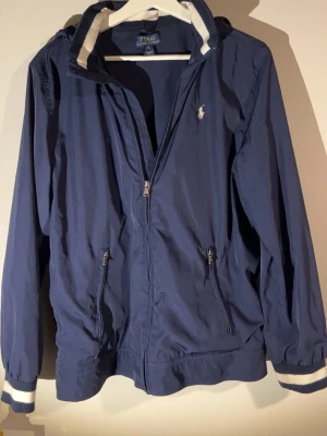 Marinblå windbreaker från Polo Ralph Lauren - Snygg marinblå vindjacka från Polo Ralph Lauren med vit logga på bröstet. Jackan har huva, dragkedja framtill, två fickor med dragkedja och ribbade muddar med vita detaljer. Perfekt för blåsiga dagar och enkel att matcha med olika outfits.                                           Hängöglan är tillbaka sydd men inget som syns!      Passformen är inte alls i XL då jag är M och den är för liten på mig!