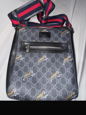 Gucci axelväska med tigerprint - Snygg axelväska från Gucci i mörkgrått med klassiskt GG-mönster och tigertryck. Väskan har en svart läderdetalj med dragkedjeficka framtill och en bred, justerbar axelrem i marinblått och rött. Perfekt för dig som vill sticka ut med en trendig accessoar.