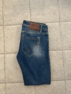 Dondup jeans  - Säljer ett par blå jeans från dondup  med klassisk femficksdesign och snygga slitningar framtill. Jeansen har en rak passform och detaljerade sömmar på bakfickan samt ett brunt läderpatch i midjan. Perfekta för en avslappnad och trendig look.