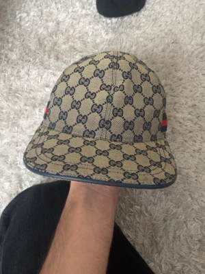 Gucci keps med GG-mönster - Snygg keps från Gucci i beige canvas med klassiskt GG-monogram i mörkblått över hela kepsen. På sidan finns en röd och marinblå rand som ger extra lyxig känsla. Perfekt accessoar för att lyfta din streetwear-look.