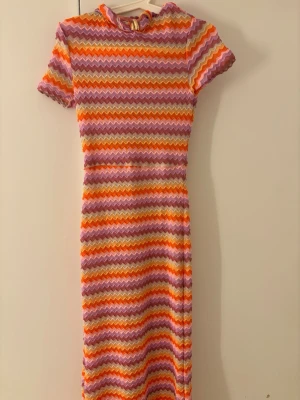 Färgglad zigzag-mönstrad klänning - Säljer en långklänning med korta ärmar och färgglatt zigzag-mönster i lila, orange, rosa och gult. Klänningen har en liten krage med guldfärgad detalj i halsen. Materialet känns stickat och stretchigt, perfekt för dig som gillar retrovibbar.