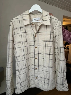 Beige rutig overshirt från Samsøe - Stilren overshirt i beige med rutigt mönster från Samsøe Samsøe. Skjortan har klassisk krage, bröstficka och knappar i trälook. Tillverkad i ett grovt, strukturerat tyg som ger en avslappnad men trendig vibe. Nypris 2000:-