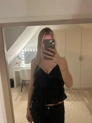 Svart volangtopp med smala axelband - Säljer en svart topp med flera volanger och smala justerbara axelband. Toppen har v-ringning och är ärmlös, perfekt för en trendig look. Materialet känns lätt och luftigt, och toppen är croppad i modellen.