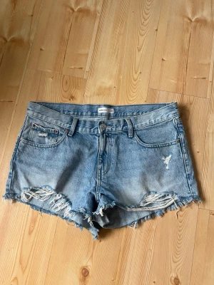 Blå jeansshorts low waist - Säljer ett par ljusblå jeansshorts från Gina tricot med råa, fransiga kanter och slitna detaljer framtill. Klassisk femficksmodell med knapp och dragkedja. Perfekta till sommaren för en avslappnad och trendig look.