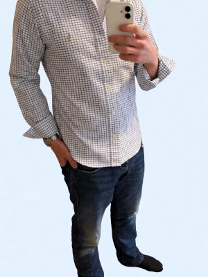 Rutig skjorta från Polo Ralph Lauren - Klassisk vit skjorta med blått rutmönster från Polo Ralph Lauren. Skjortan har knappar framtill, lång ärm och den ikoniska broderade loggan på bröstet. Tillverkad i mjuk bomull och har en normal passform. Perfekt för en clean och stilren look.