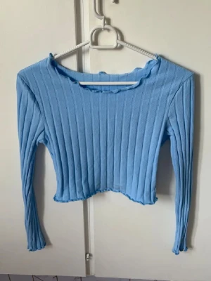 Ljusblå ribbad croppad topp - Trendig ljusblå croppad topp med långa ärmar och ribbad struktur. Toppen har en lätt vågig kant vid halsringning och nederkant, vilket ger en cool och avslappnad vibe. Perfekt att styla med jeans eller kjol för en fräsch look.