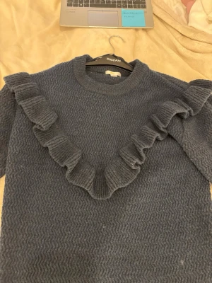 Mörkblå stickad tröja med volang - Mysig mörkblå stickad tröja från Gina tricot med volangdetalj i V-form framtill. Tröjan har rund halsringning, lång ärm och ett diskret sicksackmönster i stickningen. Perfekt för dig som gillar en trendig och bekväm look.