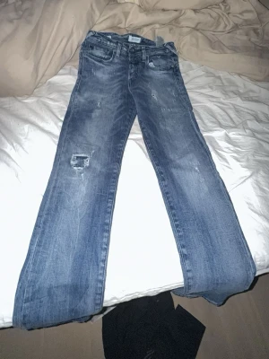 Blå bootcut jeans från Jack & Jones - Säljer ett par blå bootcut jeans från Jack & Jones med slitningar och patchdetalj på ena benet. Jeansen har klassisk femficksdesign och tydliga tvättade partier för en cool, avslappnad look. Perfekta för dig som gillar en lite retro vibe.