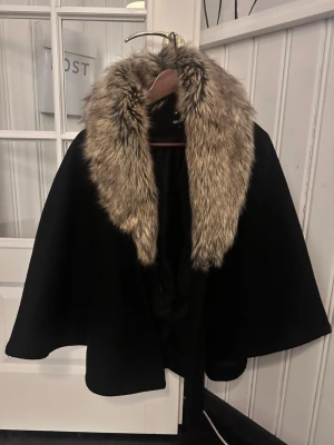 Svart cape med fuskpäls - Stilren svart cape med bred krage i beige och brun fuskpäls. Capen har en lös passform och knyts i midjan med ett band. Perfekt för dig som vill ha en elegant och trendig look med extra attityd. Från bubbleroom. Skulle säga att den passar XS-M då den är one size