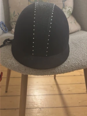 Svart ridhjälm med glitter och nitar - Snygg svart ridhjälm med glittrig mittremsa och två rader små silverfärgade nitar. Hjälmen har klassisk form, justerbart hakband och matt finish. Perfekt för dig som vill sticka ut i stallet med lite extra bling.