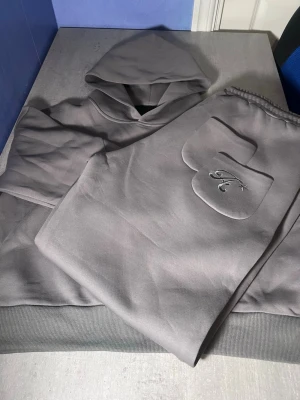 Grå hoodie med broderad ficka - Säljer en stilren grå hoodie med huva och ribbade muddar. På fickan finns ett broderat 'K' med en liten stjärna, vilket ger en unik detalj. Hoodien är i mjukt material, perfekt för chill dagar eller när du vill ha en clean look.