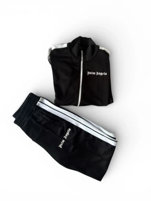 Palm Angels svart track jacket och byxor  - Svart track jacket från Palm Angels med vita ränder längs ärmarna och vit dragkedja. Jackan har  krage, två fickor med dragkedja och logga broderad på bröstet. Perfekt för en sportig och trendig streetwear-look. Jackan är storlek L (Passar M), Byxorna är storlek M