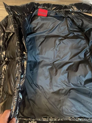 Svart pufferjacka från HUGO - Snygg svart pufferjacka från HUGO med glansig finish och hög krage. Jackan har två framfickor med dragkedja och röd HUGO-logga på bröstet. Perfekt för kalla dagar och ger en modern streetwear-look.