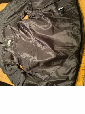 Svart pufferjacka från The North Face - Snygg svart pufferjacka från The North Face med klassisk quiltad design. Jackan har lång ärm, hög krage och är tillverkad i glansigt syntetmaterial. Perfekt för kalla dagar när du vill hålla värmen och ändå se stilren ut.