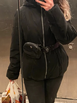  Guess pufferjacka med bälte - Äkta svart pufferjacka från Guess med huva, dragkedja framtill och ett snyggt bälte med Guess-logga och rund spänne. Jackan har en tillhörande liten väska på bältet. Perfekt för dig som gillar streetstyle och vill ha en jacka som sticker ut. Den är så snygg verkligen och är som ny inga defekter eller märken helt ny. Passar S/M. Jag tror även att jag har kvar prislappen den ser verkligen helt oanvänd ut.🖤🖤