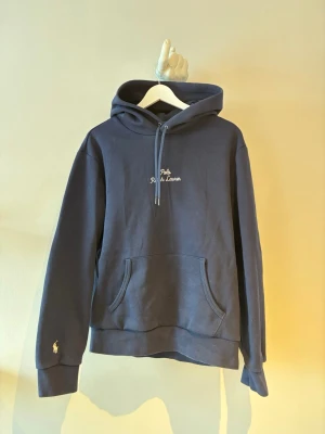 Mörkblå hoodie från Polo Ralph Lauren - Mörkblå hoodie från Polo Ralph Lauren med broderad logga på bröstet och den klassiska loggan broderad på ärmen. Tillverkad i mjuk bomullsblandning.
