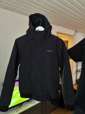 Svart vindjacka Carhartt - Snygg svart vindjacka från Carhartt med huva och dragkedja framtill. Jackan har två stora fickor, ribbade muddar och Carhartt-logga på bröstet. Tillverkad i slitstarkt syntetmaterial som står emot vind och regn. Nypris 2300:-