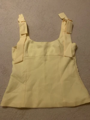 Gul linnetopp med rosetter  - Söt ljusgul linnetopp med breda axelband som knyts till rosetter. Toppen har en rak passform och ärmlös design, perfekt för varma dagar. Materialet känns mjukt och lätt, vilket gör den skön att bära. Enkel men ändå trendig detalj med knytbanden på axlarna.