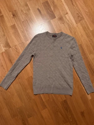 Grå kabelstickad tröja Morris - Säljer en grå/ beige kabelstickad tröja från Morris i 100% merinoull. Tröjan har rund halsringning, ribbade muddar och en liten blå logga broderad på bröstet. Liten i storleken , som en storlek S.