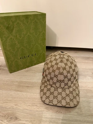 Beige GG-monogram keps från Gucci - Säljer en beige keps från Gucci med det klassiska GG-monogrammönstret över hela kepsen. Justerbar rem bak och böjd skärm. Tillverkad i canvasmaterial med detaljer i brunt. Perfekt för dig som vill ha en lyxig och trendig accessoar. Boxen följer med. Äkta!