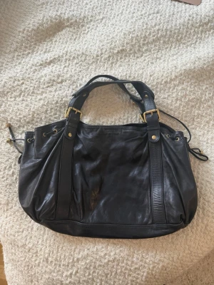 Svart handväska i skinn med detaljer - Snygg svart handväska i skinn med guldfärgade metalldetaljer och dubbla handtag. Väskan har en mjuk, lite slouchy form och dekorativa remmar framtill. Perfekt storlek för att få plats med allt du behöver. Enkel att matcha till olika outfits.